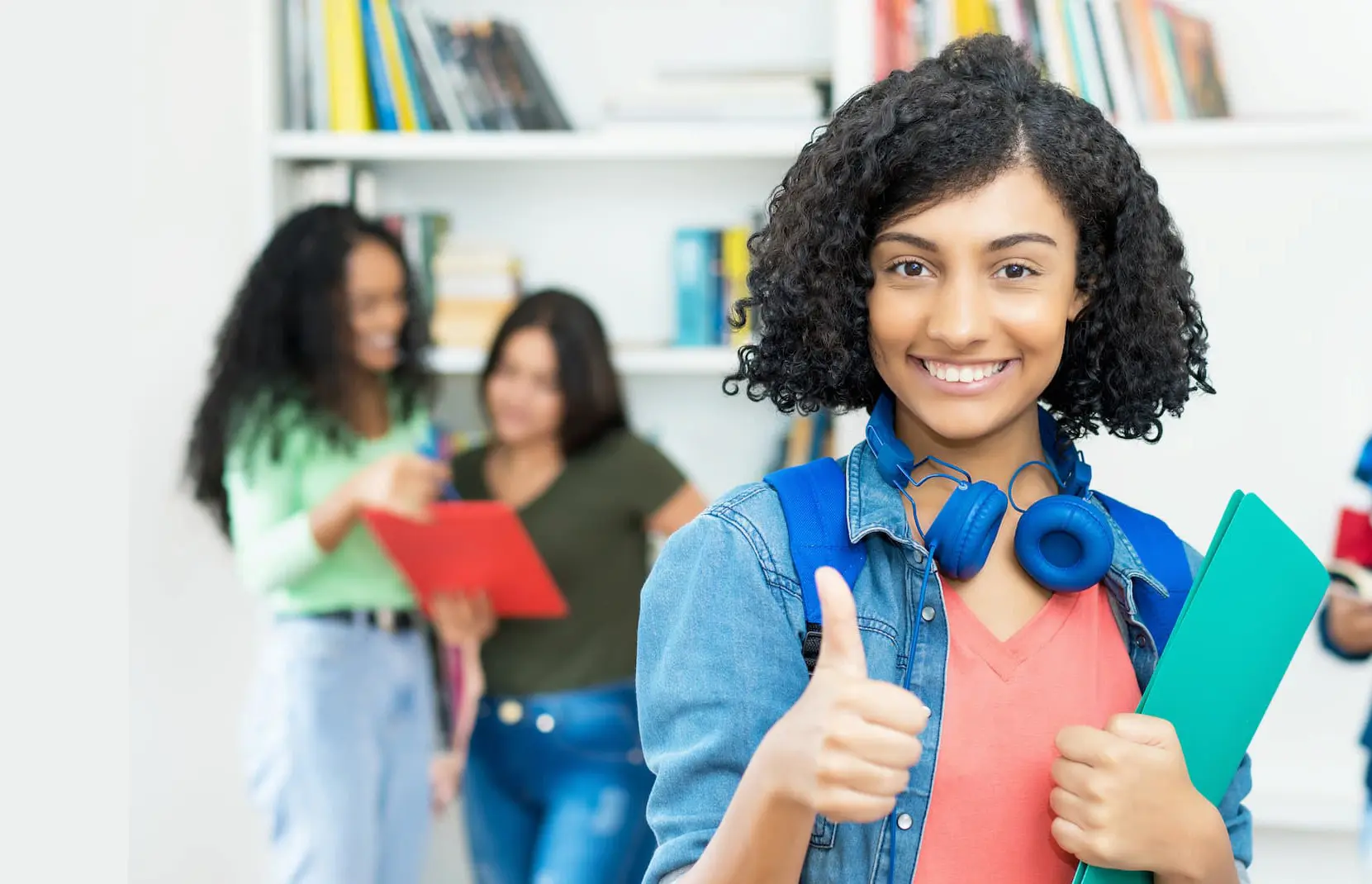 A female student holding books while smiling and giving a thumbs up, with other students in the background.