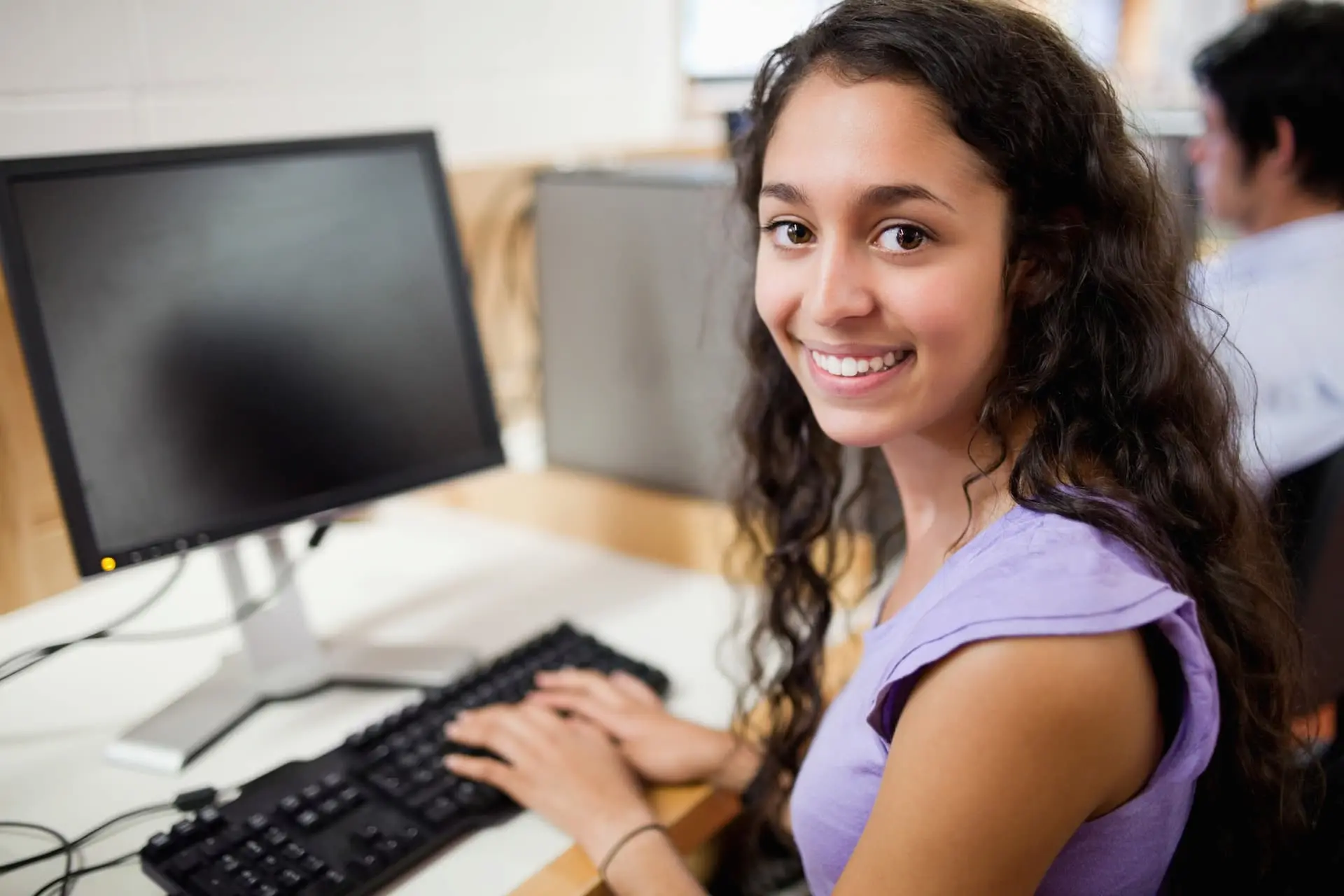 A young female student smiling, sitting at a computer.