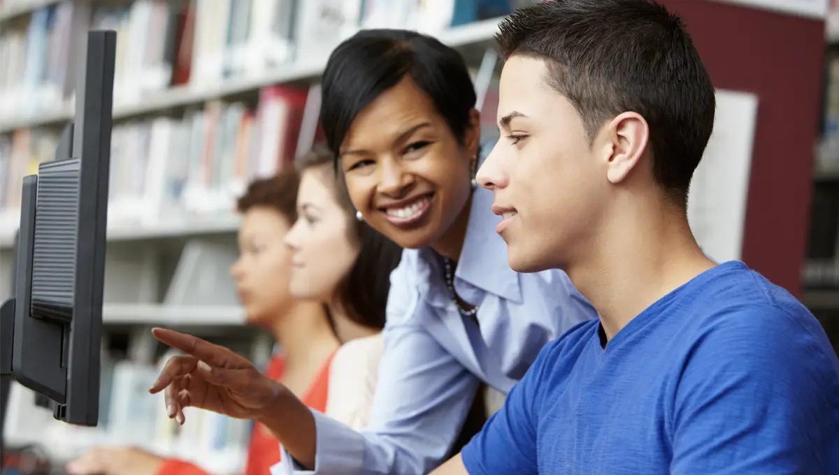 A teacher smiling with a student while looking at a monitor.