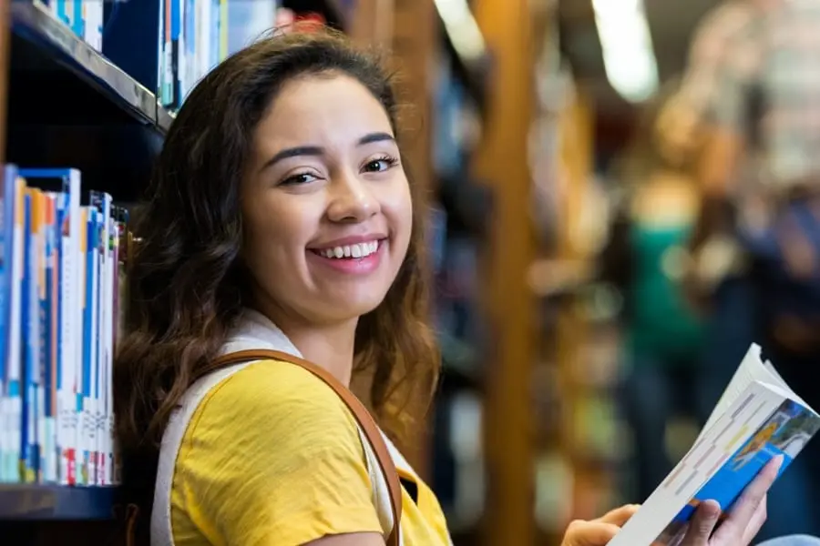 A student leaning against a shelf of books in a library, smiling.